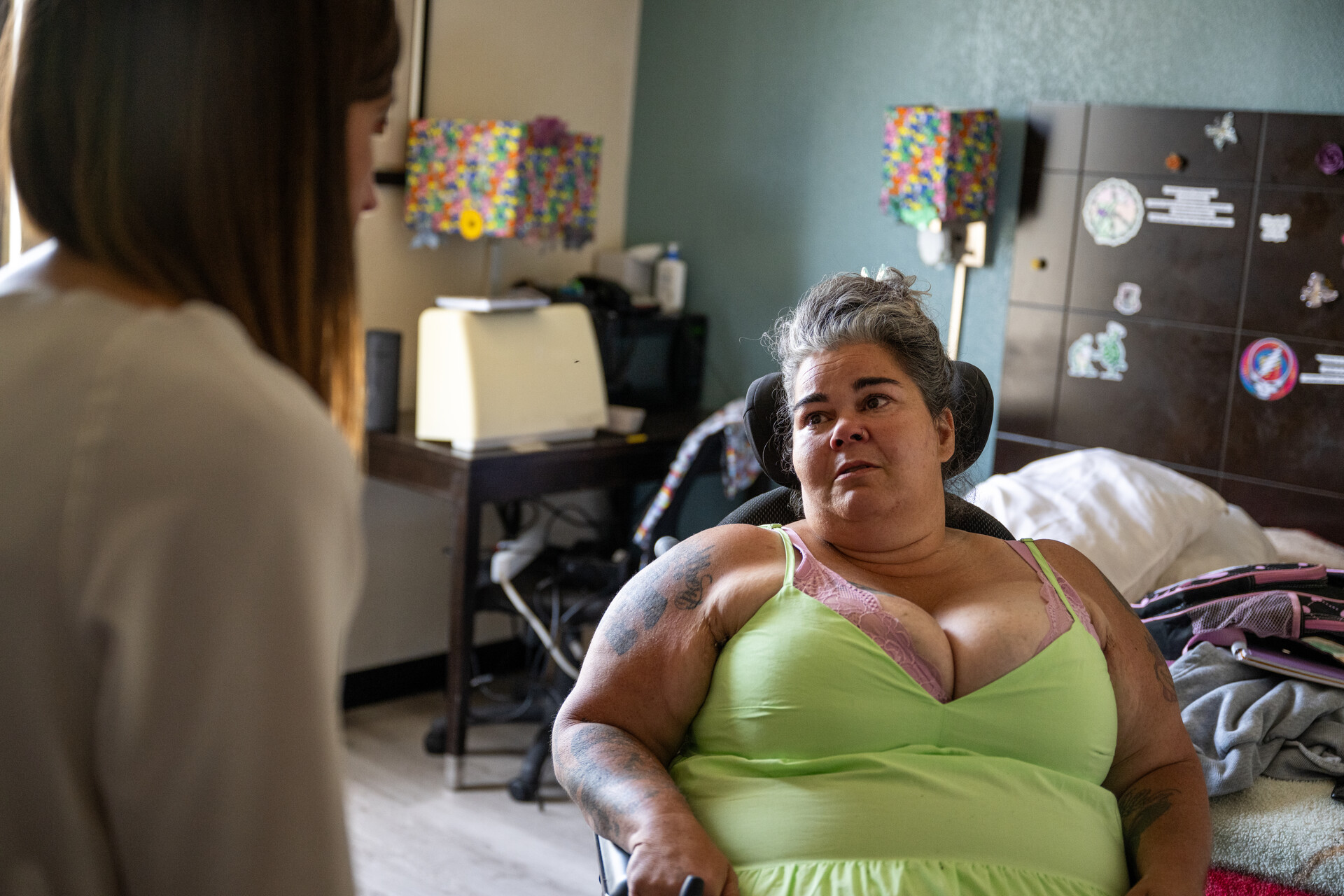 A woman seated in what appears to be a wheelchair in an apartment room with a bed in the background speaks to a woman facing away from the camera