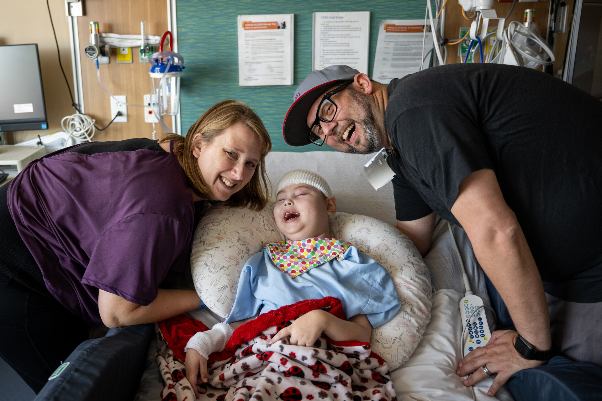 A smiling woman and man lean over a broadly smiling child who lays in a hospital bed