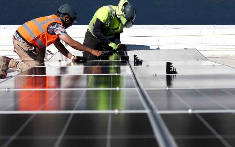 Two men wearing bright safety vests kneel over a solar panel on a rooftop.