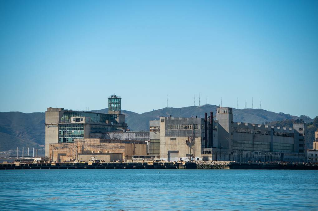 Blue bay water in the foreground with gray and tan industrial buildings along the bay's edge in the background. In the distance are hills and above is a blue sky.