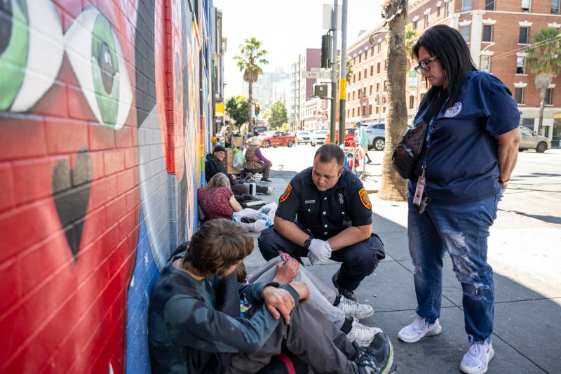 A male paramedic crouches down in front of two people sitting on the street, leaning against a wall with a mural, as a middle-aged woman stands over them.