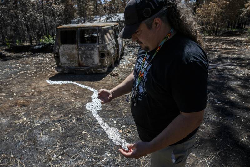 A man with long hair flowing out from beneath a hat holds a piece of burned metal in his hands. In the background, a charred vehicle.