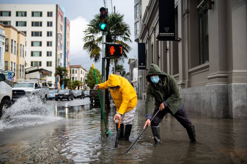 Two people in rain gear stand on a flooded San Francisco street, using rods to unclog a storm drain.