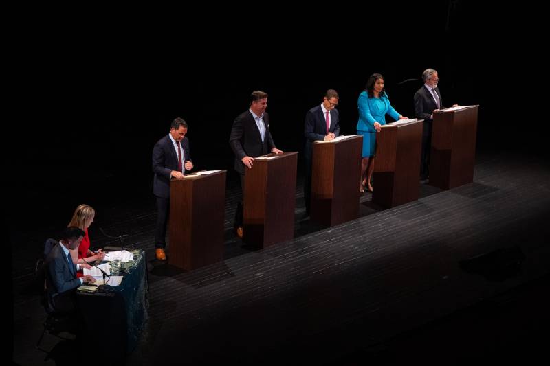 Four men and one woman stand behind podiums on a dark stage.