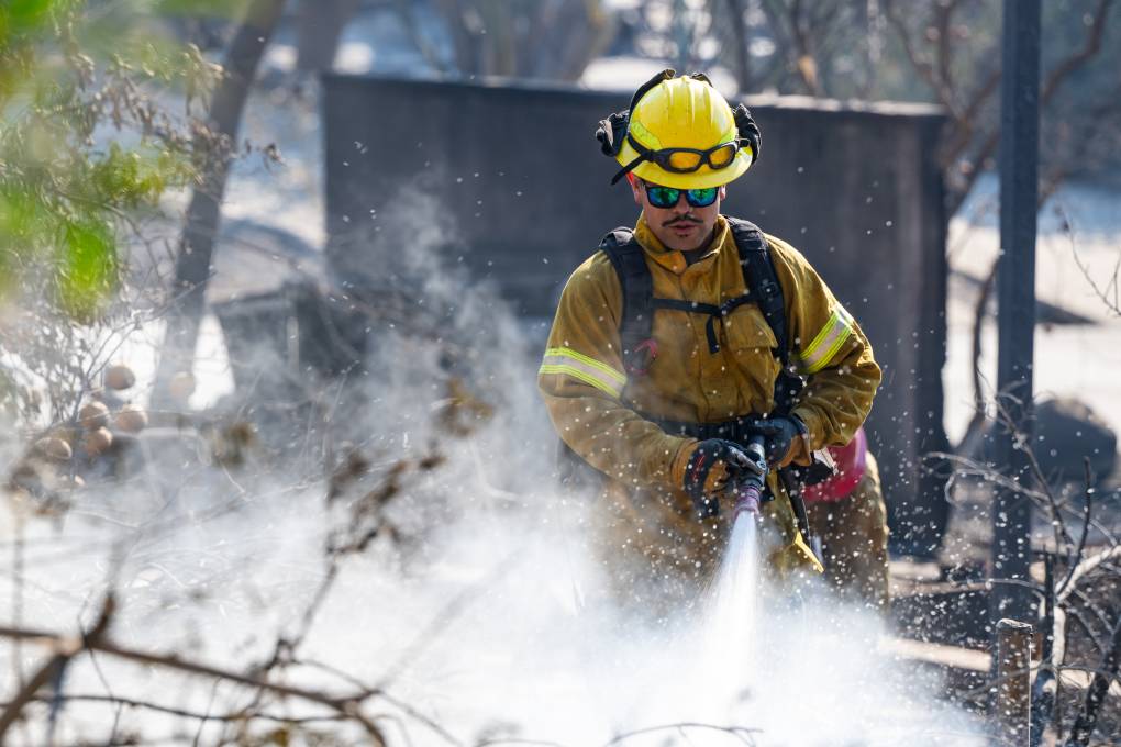 A firefighter points a hose to the ground amid smoldering ruins.