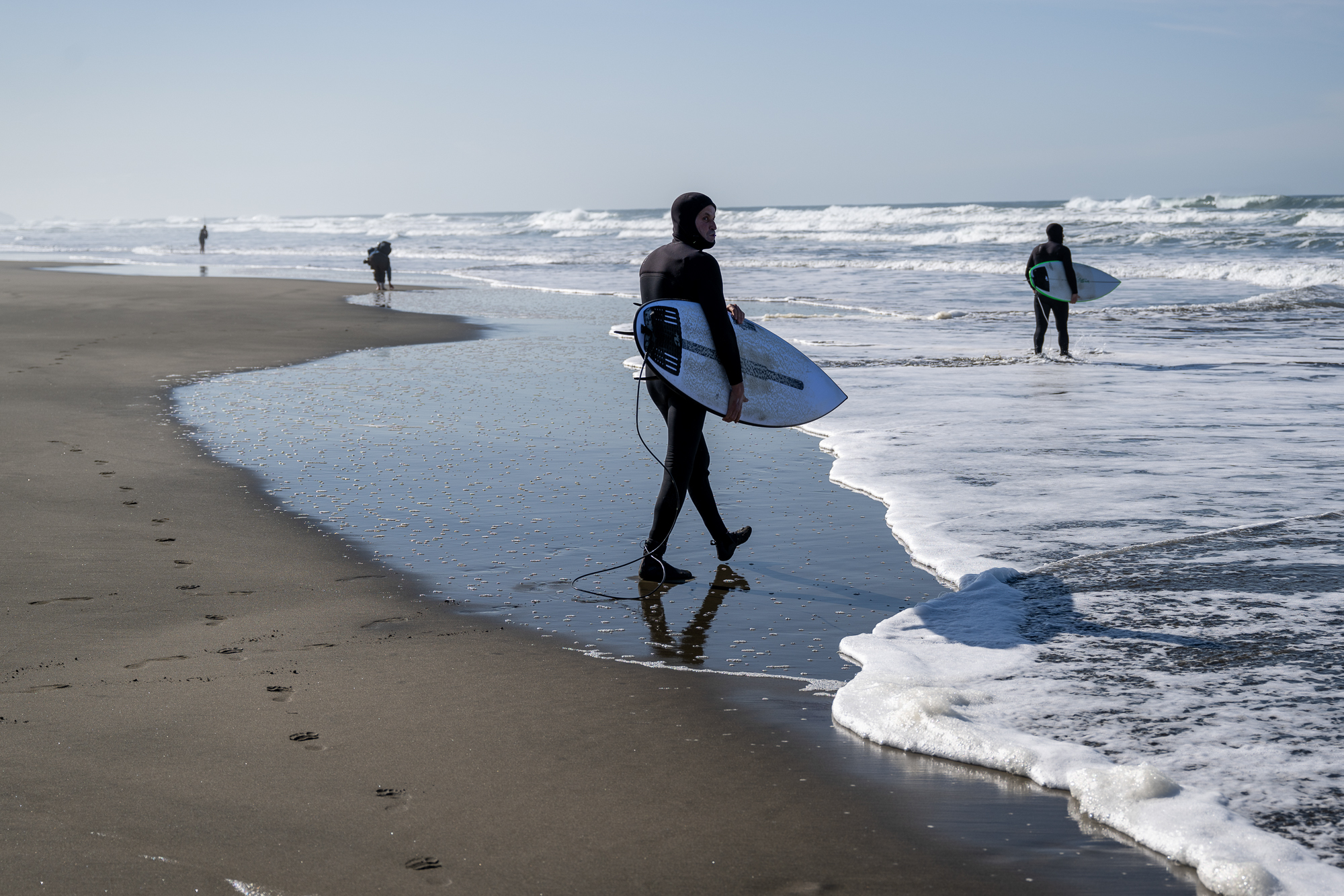 A person in a full body wetsuit, with hood, holding a surfboard under his arm, walks into the ocean.