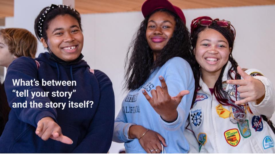 Three smiling students pose together and gesture toward the camera. White text over the image asks, “What’s between ‘tell your story’ and the story itself?”