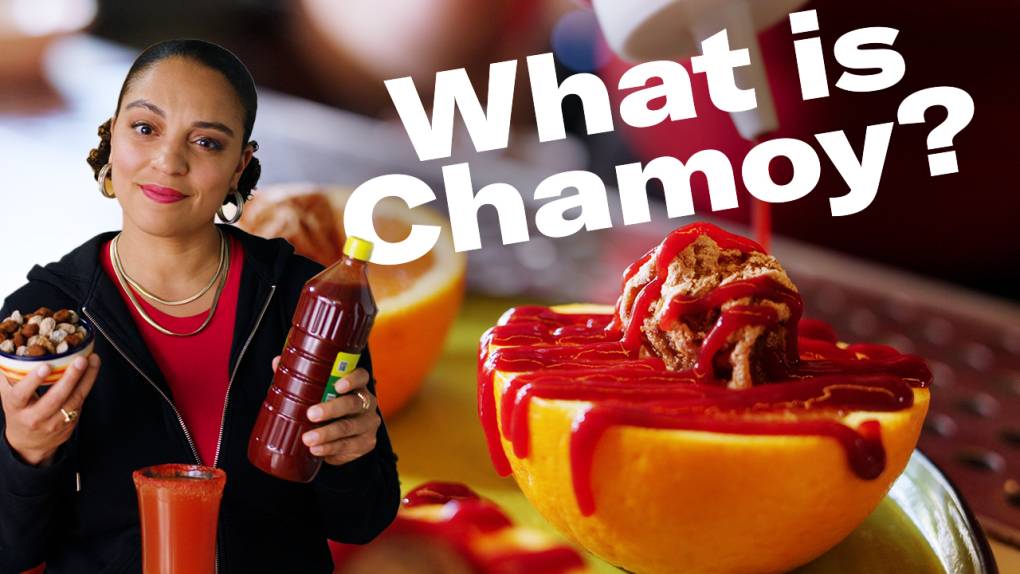 A young light-skinned black woman stands in the foreground holding a bottle of chamoy. In the background, a large bottle of chamoy sauce is squeezed onto half an orange.