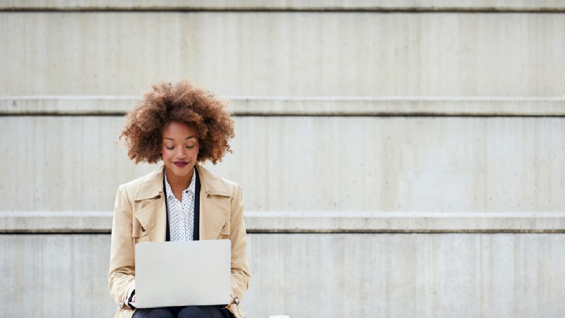 Young businesswoman using laptop while sitting on steps. Full length of female professional working on staircase. She is wearing long winter coat.