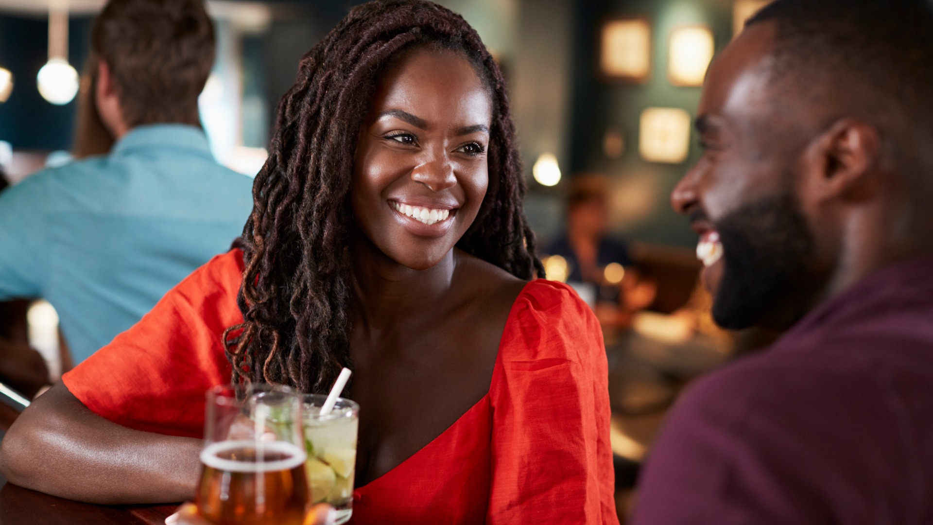 A woman in red smiles at her date while holding a cocktail at a bar.