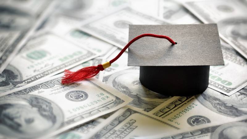 A graduation cap with a red tassel sits on top of a layer of one hundred dollar bills.