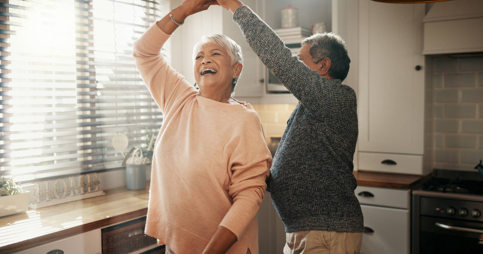 Image depicts an older couple laughing, dancing together in a kitchen.