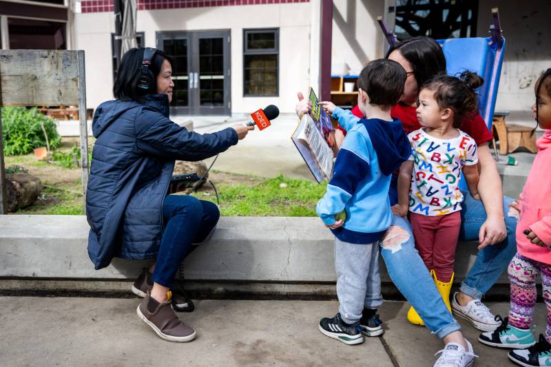 Reporter sits on the edge of a garden bed and interviews preschool aged children with a KQED microphone.