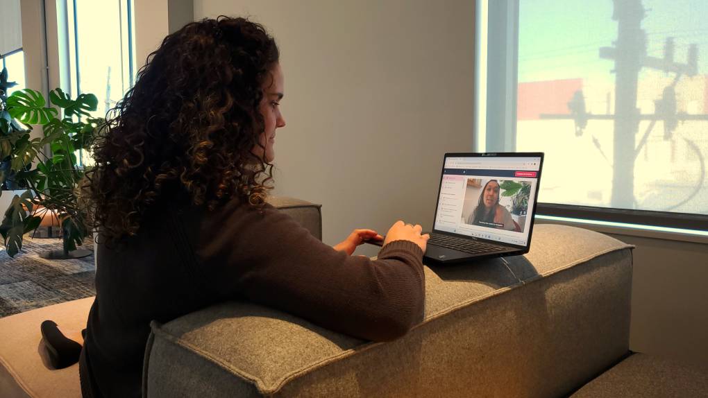 A woman sits comfortably on a couch with a laptop and watches another woman speaking in an instructional video.