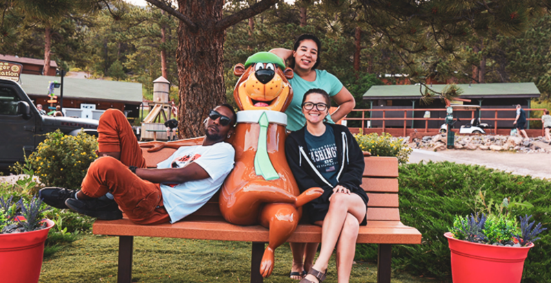 Three young people in their 20s pose on a bench with a statue of Yogi Bear smiling in the center.