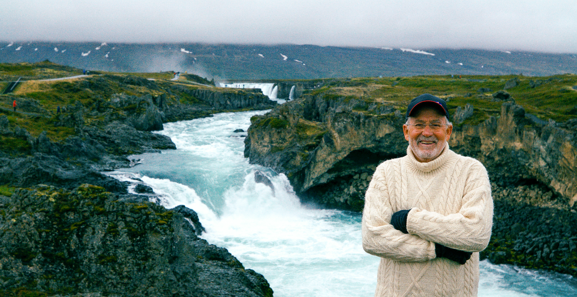 Host Joseph Rosendo stands in front of a waterfall in Iceland, wearing a sweater and baseball cap.