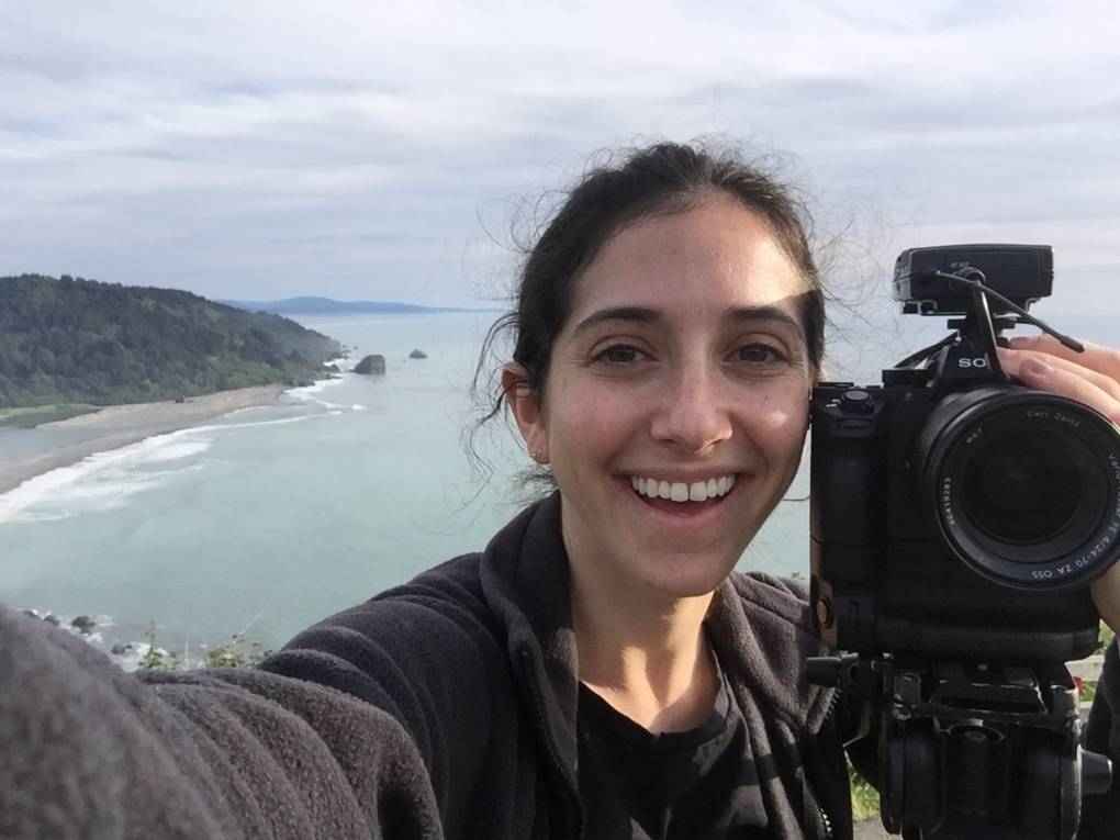 Margaret Katcher seen smiling and holding a video camera with the California coast line in the background.