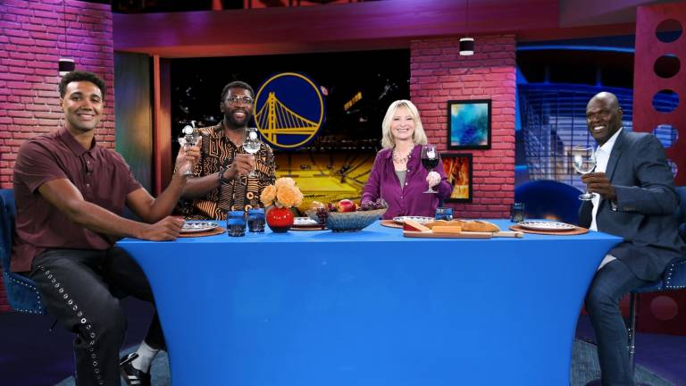 Photo of the Check, Please! Bay Area set. Center is a dinner table with the folliwing panelists seated facing the camera from left to right: Current Golden State Warrior Trayce Jackson-Davis, former Warrior Festus Ezili, host Leslie Sbrocco, and former Warrior Adonal Foyle. Each is toasting with a wine glass.