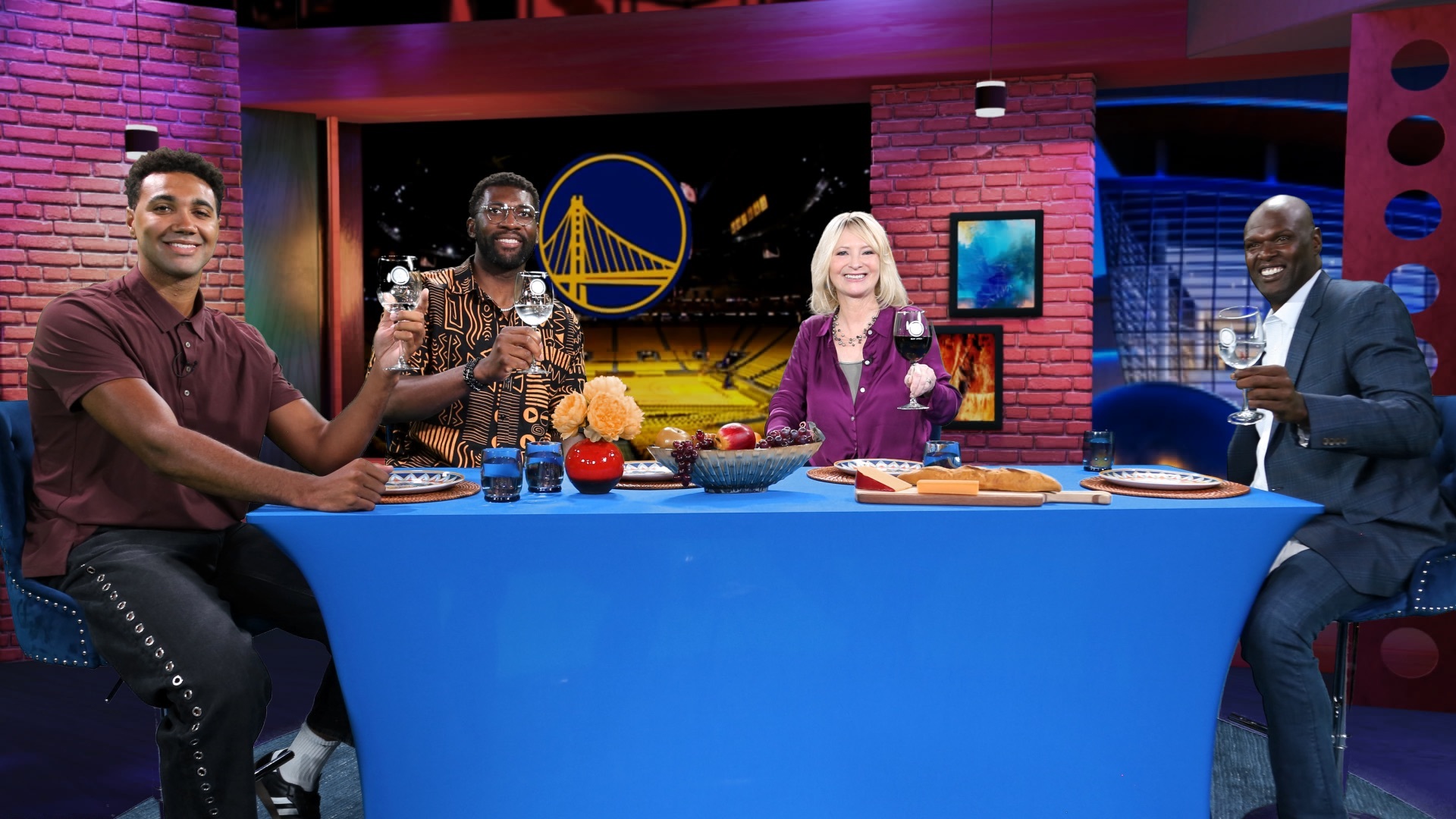 Photo of the Check, Please! Bay Area set. Center is a dinner table with the folliwing panelists seated facing the camera from left to right: Current Golden State Warrior Trayce Jackson-Davis, former Warrior Festus Ezili, host Leslie Sbrocco, and former Warrior Adonal Foyle. Each is toasting with a wine glass.