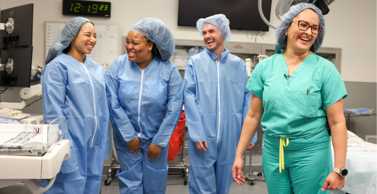 Four people dressed in scrubs stand laughing in a hospital operating room.