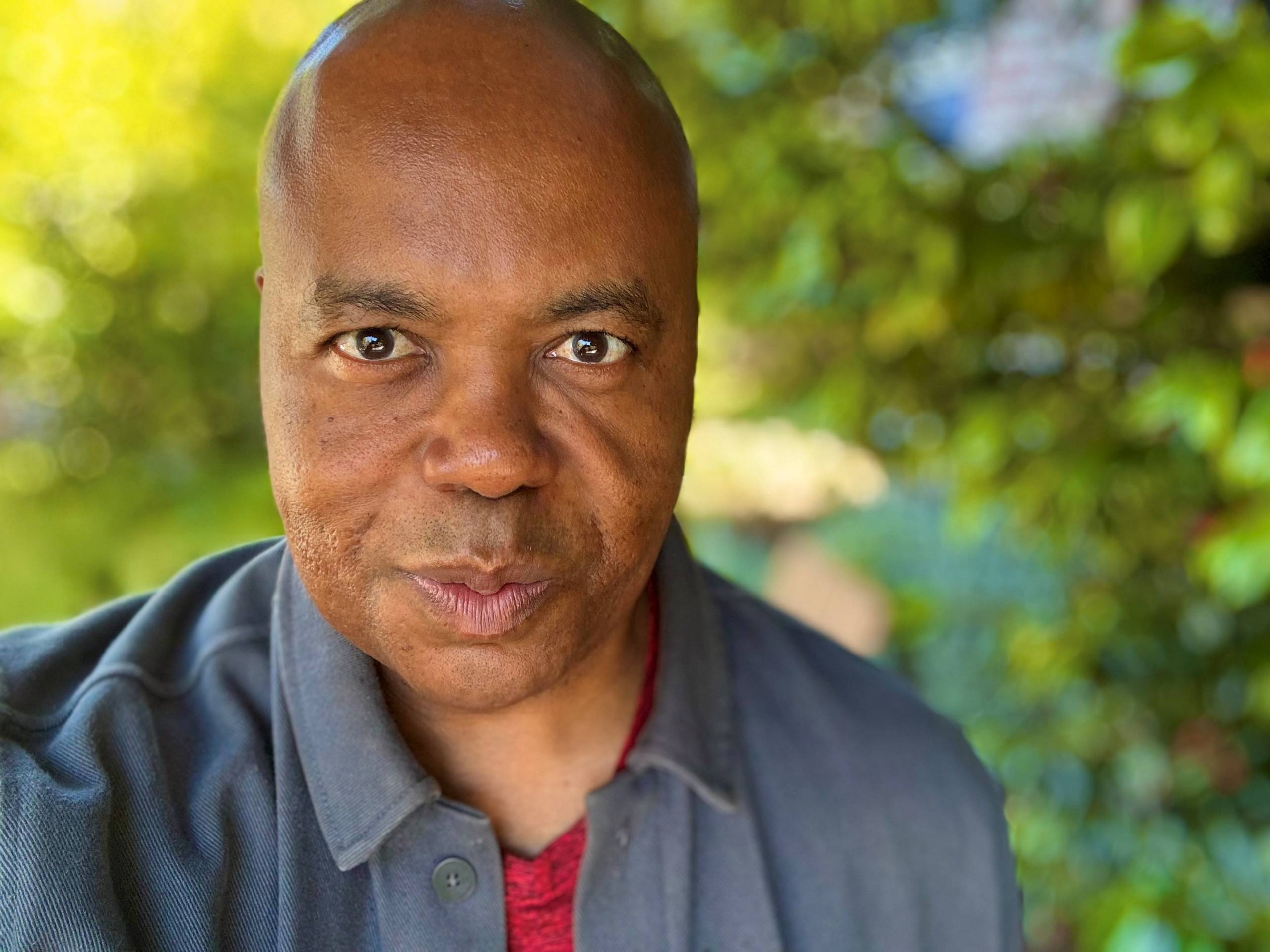 A headshot of Glynn Washington, host and co-founder of Snap Judgment. Glynn's face is in the foreground. Hist expression is friendly, with lips closed. He is wearing a blue, button down shirt with the top part of a red undershirt exposed under the neck. In the background are leafy green trees.