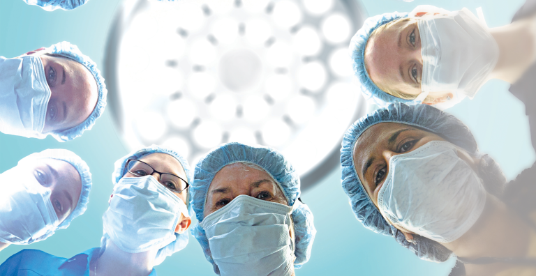 Six women in in scrubs and face masks look down at the camera in an operating room with a light above them.
