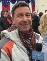 A candid close-up of KQED reporter Scott Shafer smiling at the 2017 Donald Trump Presidential inauguration, wearing winter gear and holding a KQED microphone.