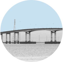 A span of the San Mateo Bridge is shown above still bay waters. The image is in black and white and contained in a light blue circle.