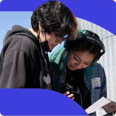 two students looking down at a piece of paper, one holding a microphone and wearing headphones